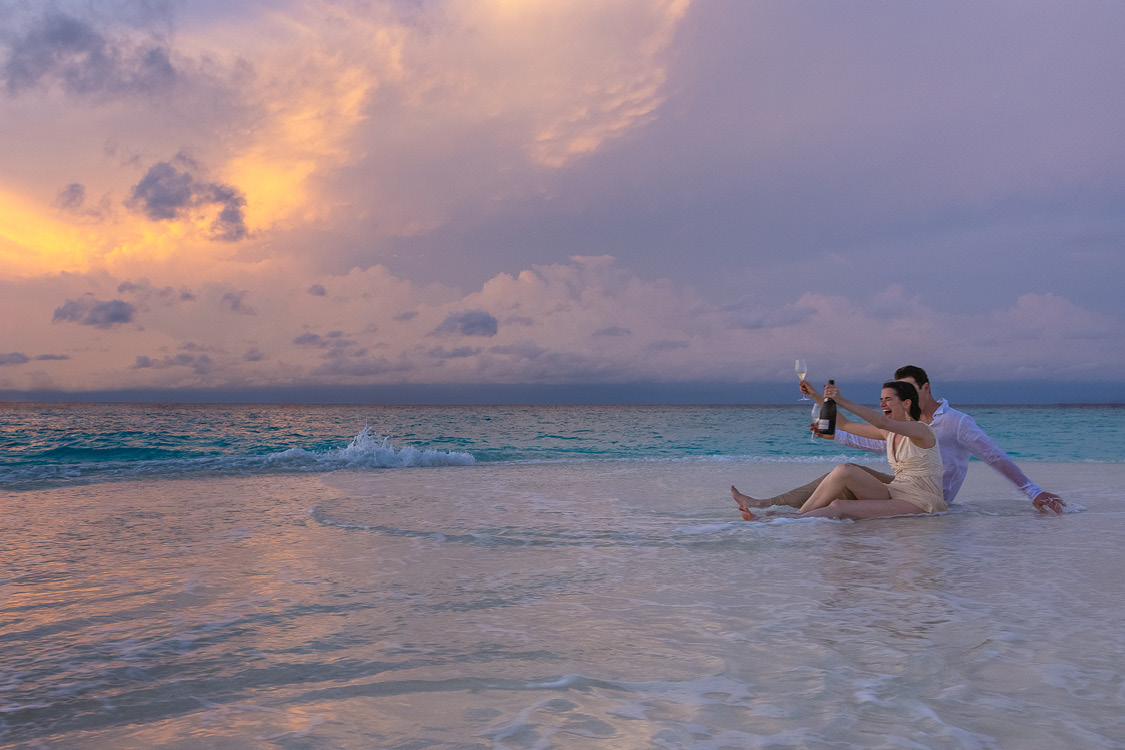 An excited couple toasting with champagne while sitting on a sandbank at sunset