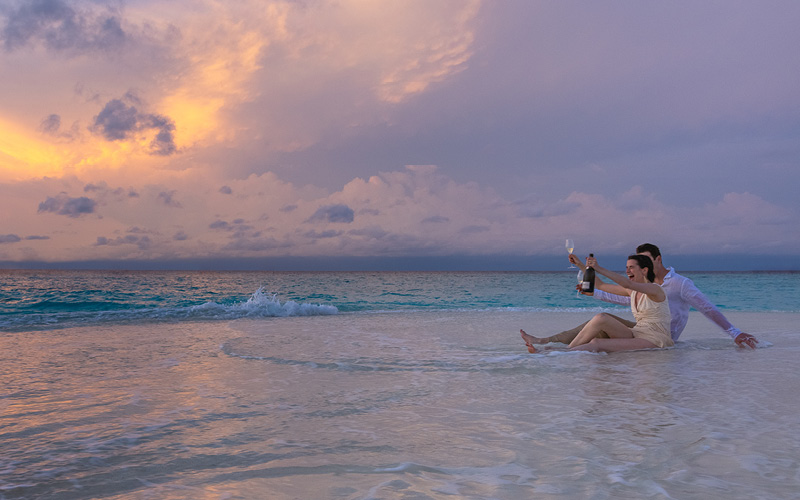 An excited couple toasting with champagne while sitting on a sandbank at sunset
