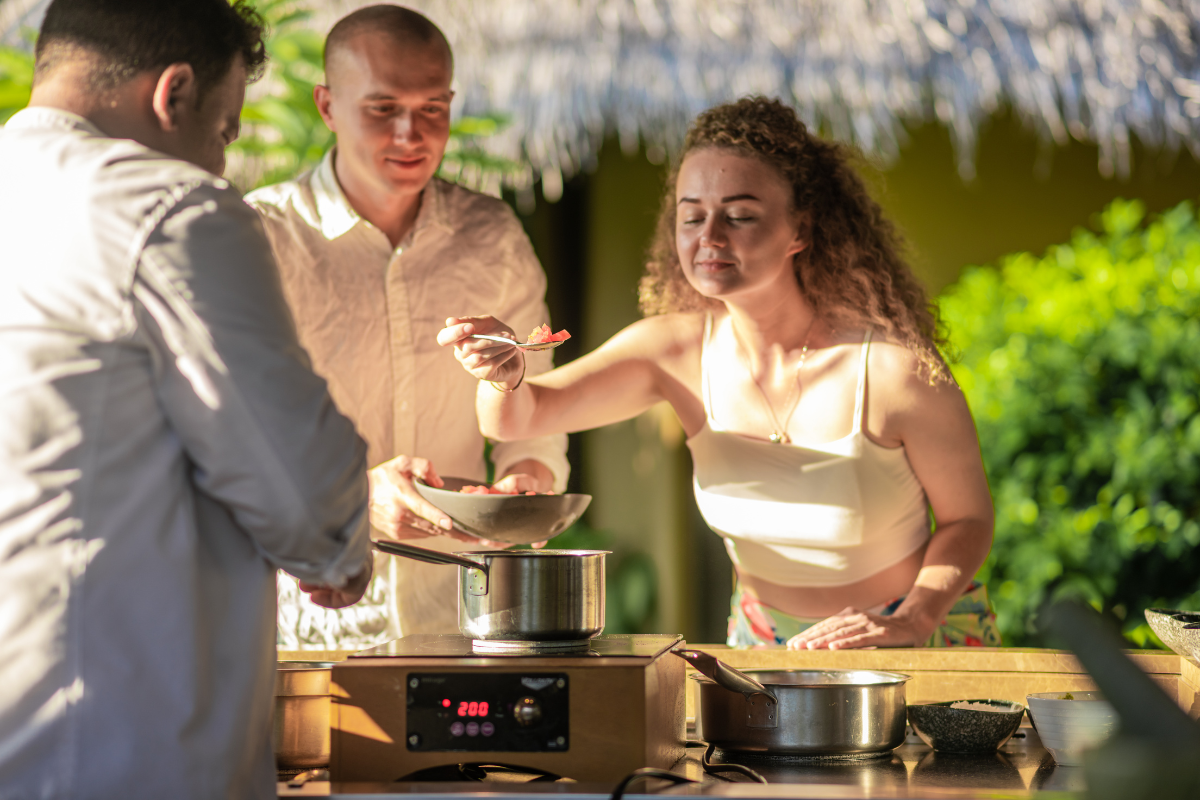 A woman holding up a spoon in front of a chef, savouring the aroma