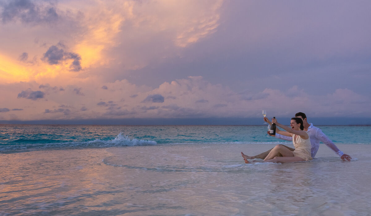 An excited couple toasting with champagne while sitting on a sandbank at sunset