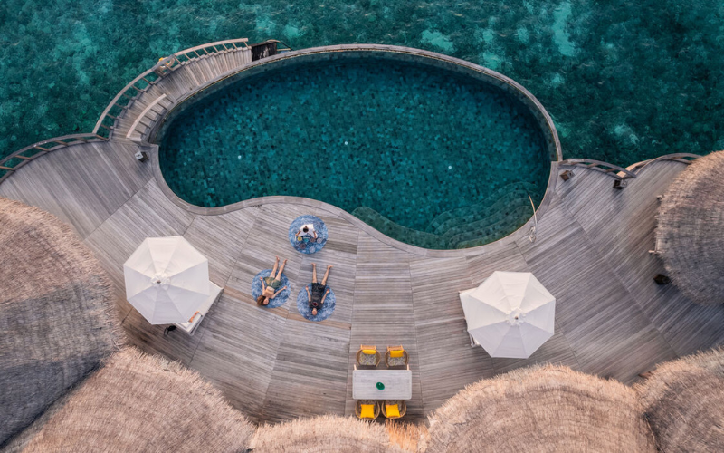 Aerial image of two people and a yoga instructor practicing yoga on a deck of an over water villa with private pool at The Nautilus Maldives