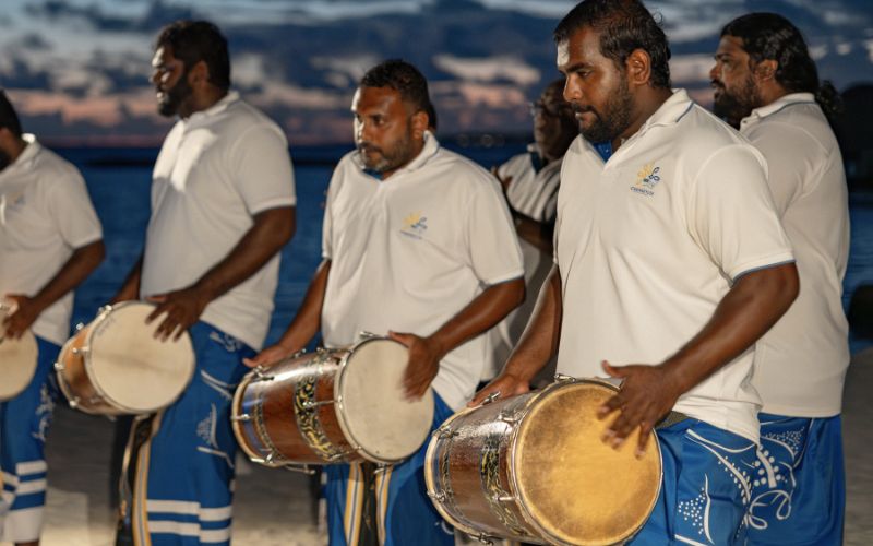 Group of Boduberu drummers dressed in traditional attire, performing near the beach at dusk. The musicians are holding large drums and wearing white shirts and blue sarongs with decorative patterns. The sky is darkening, creating a dramatic backdrop for the rhythmic and energetic performance.
