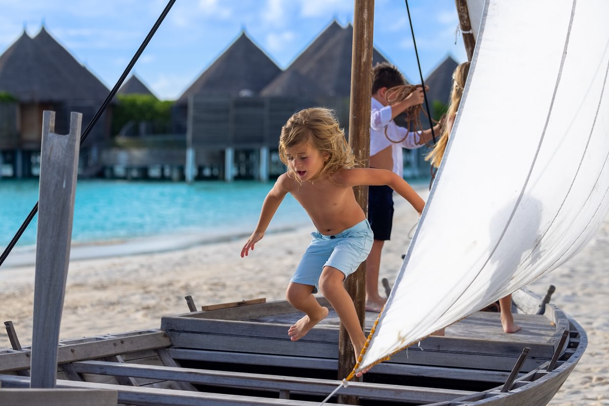 Children enjoying a beachside adventure on a traditional wooden boat, with water villas in the background.